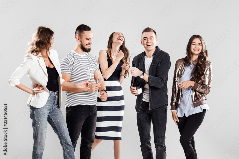 Obraz premium Company of friends in fashionable casual clothes stand together and smile. Guy opens a bottle of champagne
