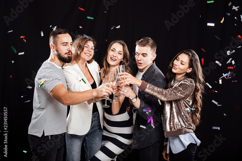 Company of friends in stylish casual clothes stand together and clink glasses with champagne on a black background and confiture around. Party time