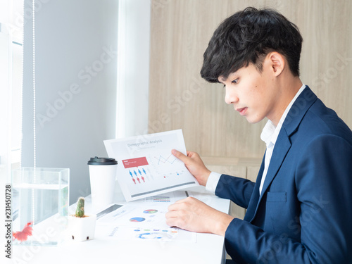 the young asian business man hold the chart during working, he has the coffee cup, fish, chart and cactus on the table