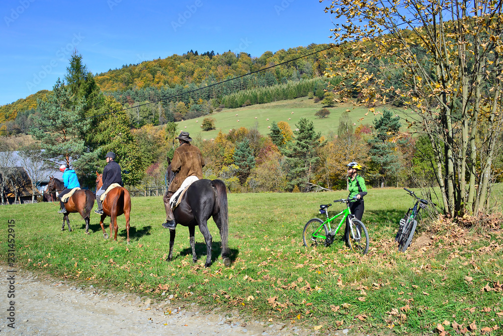 Woman next to a green bicycle looking at a three people riding horses in countryside nature tour in Low Beskid (Beskid Niski) in autumn sunny day. Woman next to a green bicycle looking at a three people riding horses in countryside nature tour in Low Beskid (Beskid Niski) in autumn sunny day.