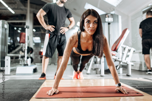 Fototapeta Naklejka Na Ścianę i Meble -  Beautiful athletic girl dressed in black sports top and tights is doing plank under the supervision of a coach in the gym