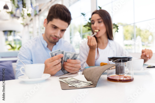Displeased confused loving couple sitting in cafe holding check and money.