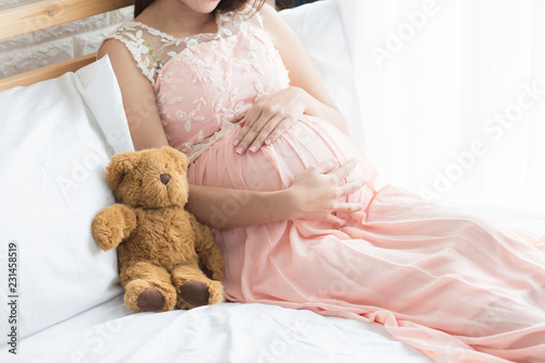 Pregnancy woman in pink dress and teddy bear on the bed in bedroom.