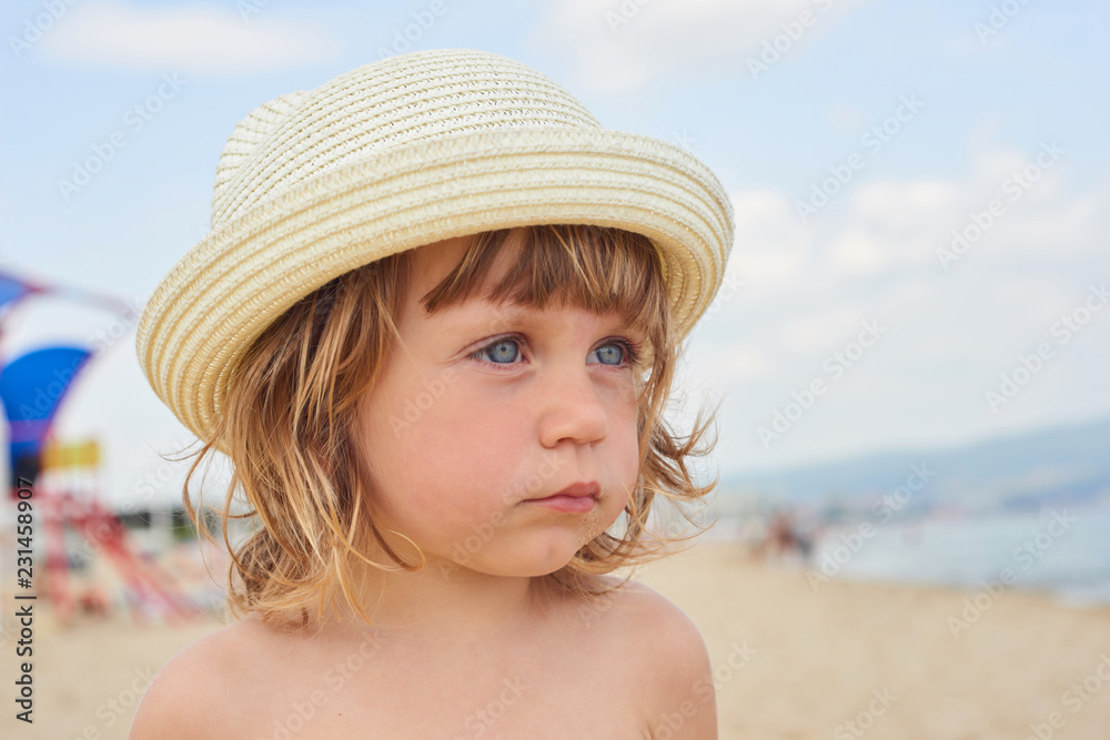 Little two year old girl with blue eyes in a bright wicker hat, standing on the beach near the sea