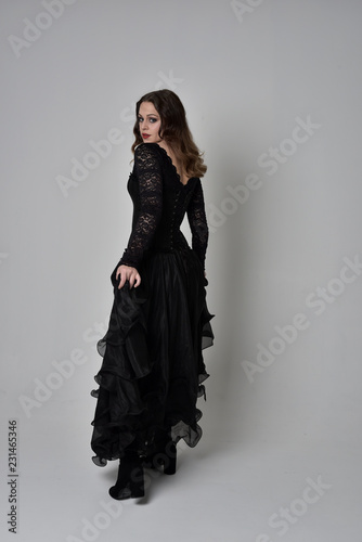 full length portrait of brunette girl wearing long black lace gown with corset.  standing pose with back to the camera, grey studio background.
