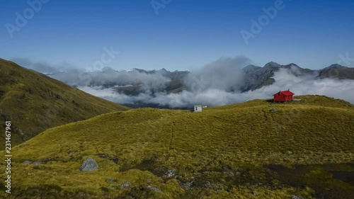 Beautiful scenery in New Zealand with red mountain hut on sunny summer day with clouds and fog down in the deep valley. Brewster hut, Haast Pass. Timelapse video.