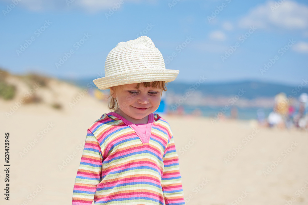 Little two year old girl with blue eyes in a bright wicker hat, standing on the beach near the sea