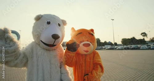 Two cute growth dolls of bears standing outside on the big modern buildinf and parking background and waving their hands or paws. Camera zooming out. photo