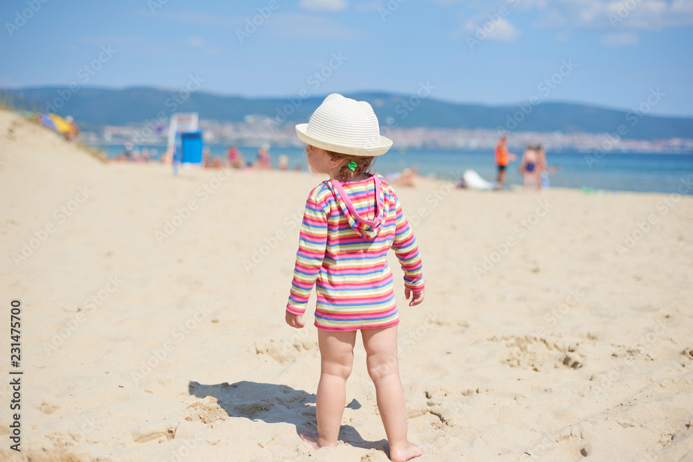 Little two year old girl with blue eyes in a bright wicker hat, standing on the beach near the sea