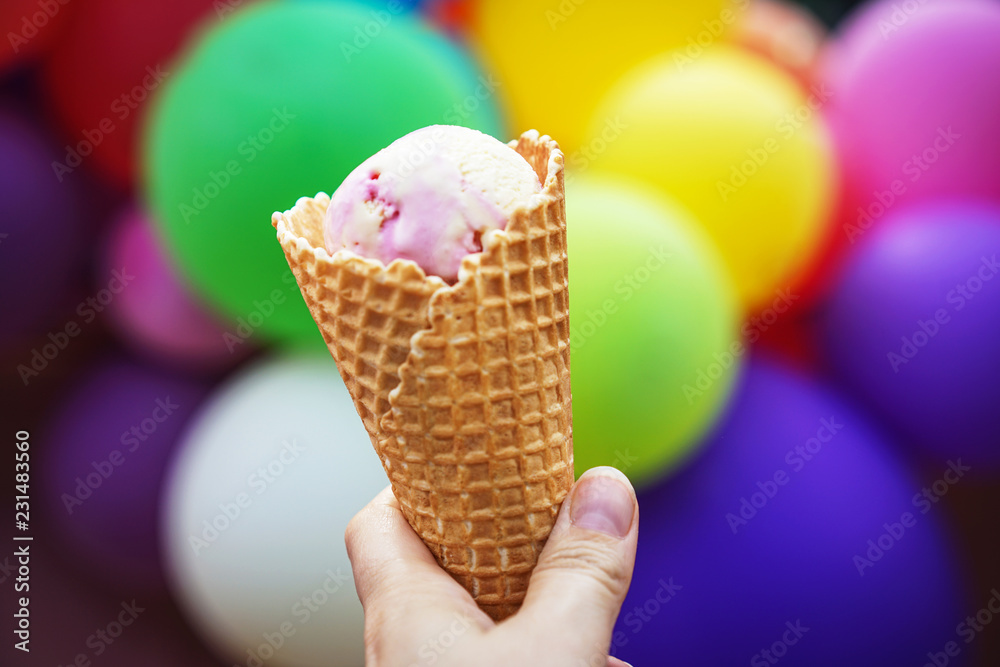 Close up of woman hand holding fresh waffle cone with strawberry ice cream on the background of colorful balloons Close up of woman hand holding fresh waffle cone with strawberry ice cream on the background of colorful balloons