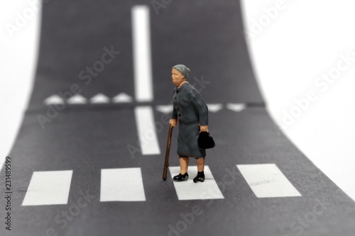 persona anziana in miniatura che attraversa la strada sulle strisce pedonali aiutandosi col bastone photo