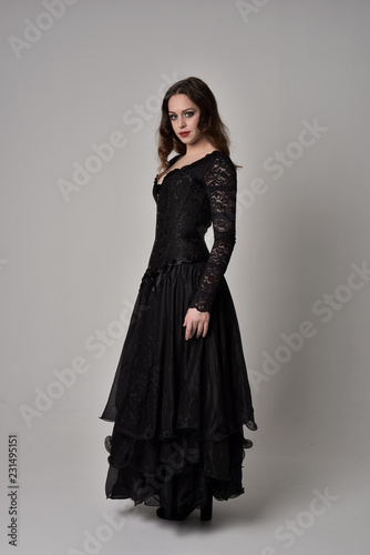 full length portrait of brunette girl wearing long black gown with corset. standing pose on grey studio background.