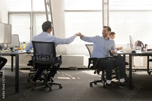Friendly male coworker buddies fist bumping at work celebrate good teamwork result, happy smiling colleagues share success motivated by great relations, corporate culture help support respect concept