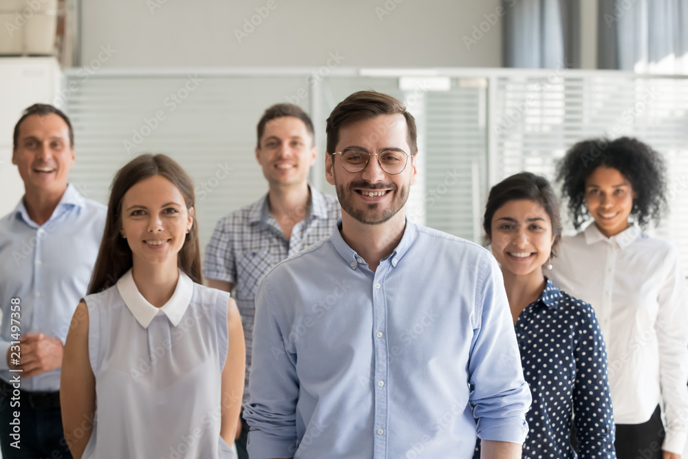 Smiling leader ceo or professional business coach looking at camera posing in office with diverse happy team at background, successful startup founder, corporate employee with staff members portrait