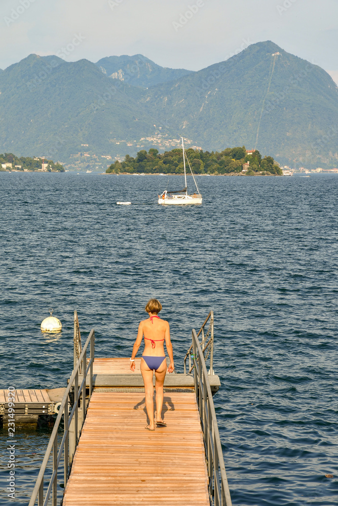 Young woman from behind walking on a wooden pier at Maggiore Lake with Madre Isle and mountainous coast in the background, Baveno, Piedmont, Italy Young woman from behind walking on a wooden pier at Maggiore Lake with Madre Isle and mountainous coast in the background, Baveno, Piedmont, Italy