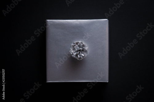 Square shape gift wrapped in silver paper, decorated with silver pine cone on black background