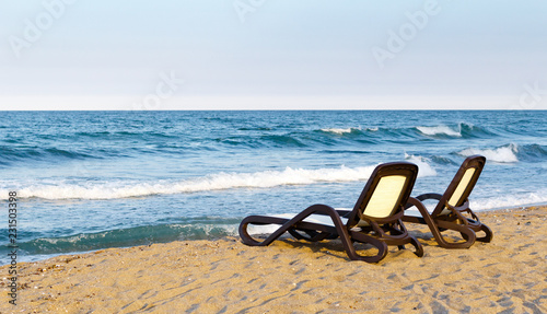 Lonely pair of beach deckchairs on seashore in front of sea, sunset.