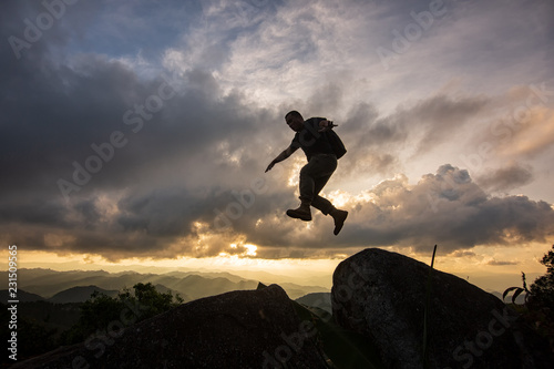 Silhouette Freedom-young man is jumping over precipice between two rocky mountains at sunset.
