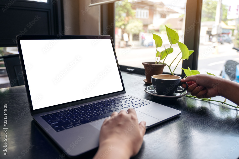 Fototapeta premium Mockup image of a woman's hands using laptop with blank white desktop screen while drinking hot coffee on wooden table in cafe