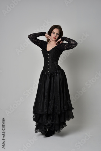 full length portrait of brunette girl wearing long black gown with corset. standing pose on grey studio background.