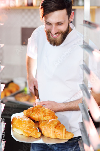 Handsome baker in white uniform holding in his hands a tray full of freshly baked croissants against the background of a bakery