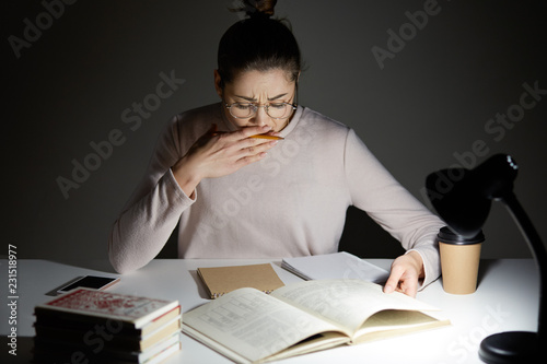 People and tiredness concept. Sleepy student yawns, poses at desktop, work overtime, during night, prepares for upcoming exam, looks at book, sits near reading lamp, drinks aromatic beverage photo