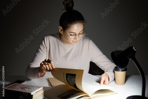 Photo of serious female sudent turns over pages in book, searches necessary information for writing course paper, wears round big spectacles, sits at table with notebook and textbooks, drinks coffee photo