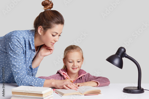 Horizontal shot of serious young mother teaches her small light haired daughter to read, shows something in book, pose at desktop with literature and readinglamp, isolated over white background photo