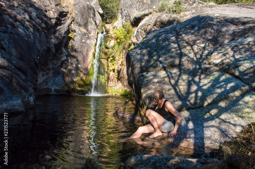 Caucasian woman sitting on a big stone, in the summer, near a little lake with a waterfall with her feet in the water