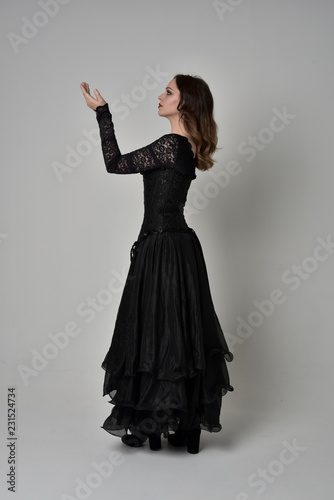 full length portrait of brunette girl wearing long black gown with corset. standing pose on grey studio background.