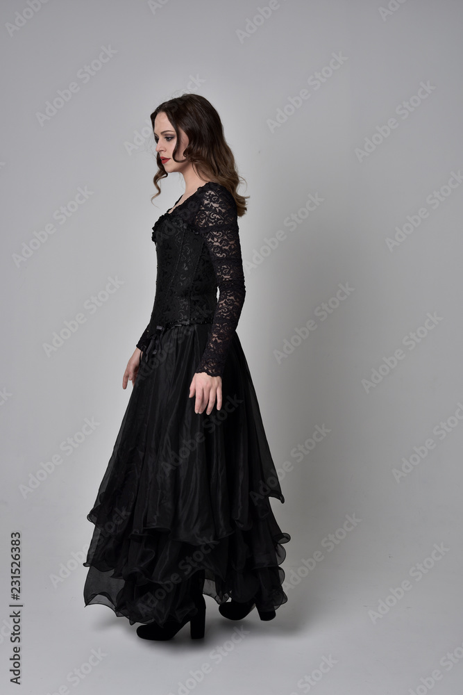 full length portrait of brunette girl wearing long black gown with corset. standing pose on grey studio background.