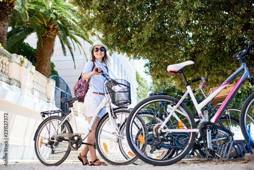 Young happy woman in sunglasses, white shorts, blouse and hat with a bicycle smiling to the camera in shadow of palm trees at bike parking on bright sunny day. Active lifestyle and vacations concept.