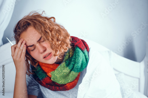 Portrait of exhausted young girl in woolen scarf arounf her neck feeling unwell. Sick girl in coloured scarf touches her forehead. photo