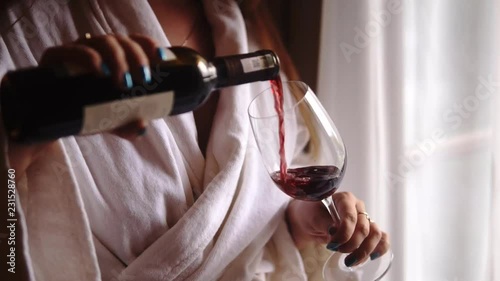 Young woman pours bottle of red wine into a glass. Girl staying near the balcony window dressed in a white hotel bathrobe. Luxury hotel resort spa conception.
