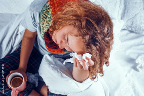 Exhauted woman with a cup of tea rests in bed because of sickness. Unhealtly young girl in scarf around neck holds a tissues pressed to her forehead. photo