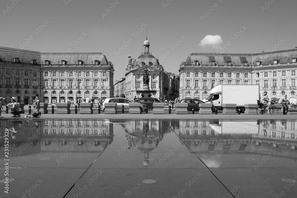 Black and white picture of Place de la Bourse, a square in Bordeaux France with a reflection in the water. People are walking, trucks and cars are riding
