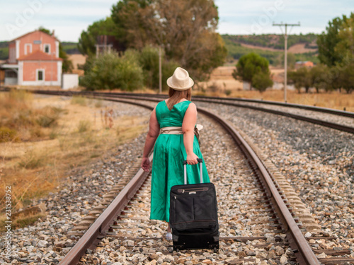 A blonde woman with a green dress and a straw hat walking on the train tracks carrying a suitcase in autumn
