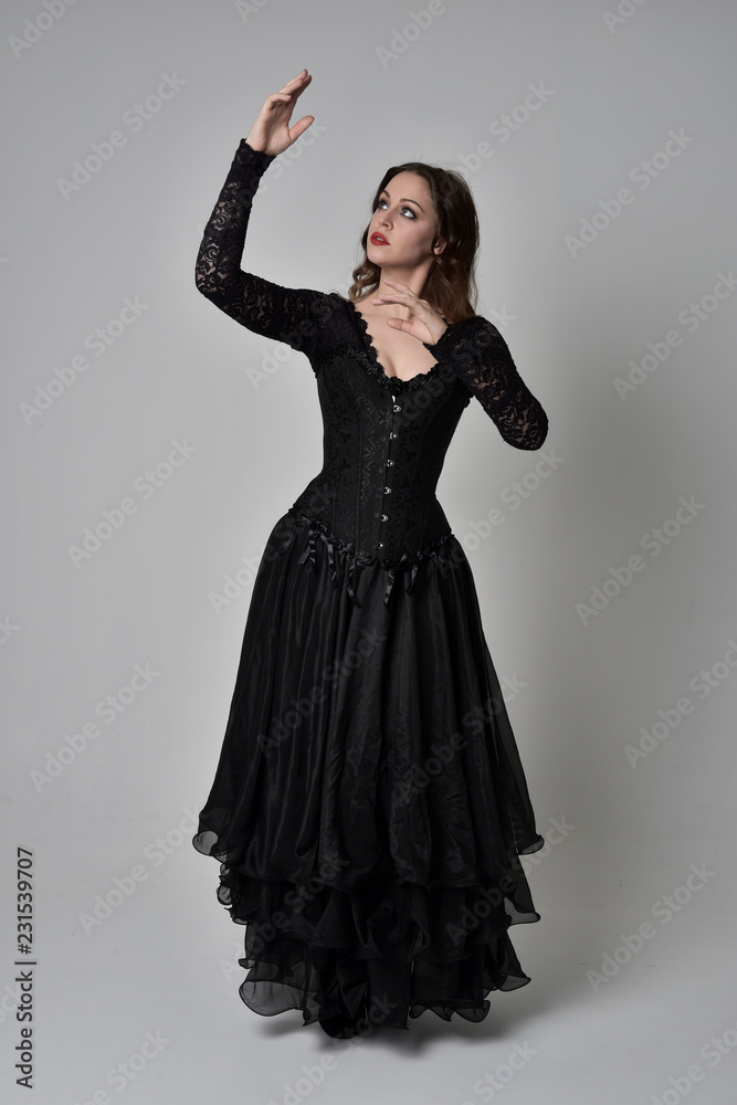 full length portrait of brunette girl wearing long black gown with corset. standing pose on grey studio background.