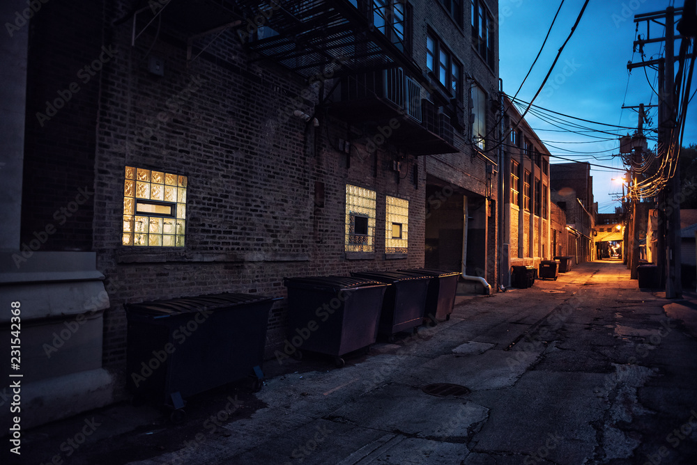 Dark and scary downtown urban city street alley scene with an eerie vintage industrial warehouse factory with illuminated windows at night