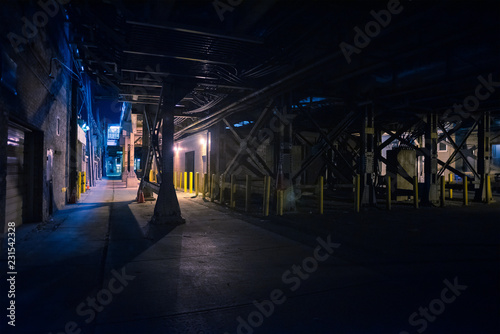 Dark and scary downtown urban city street alley under an eerie and illuminated vintage industrial railroad subway station bridge at night