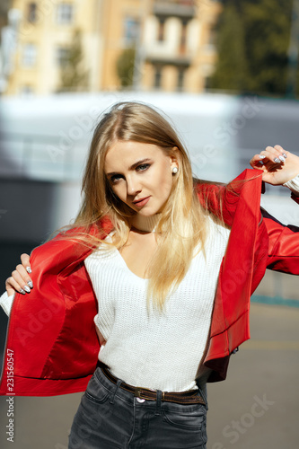 Luxurious blonde model with long hair wearing red leather jacket posing in sun light