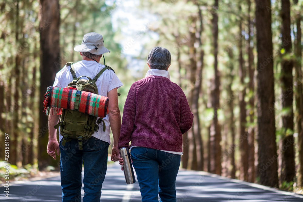 Couple of old aged senior traveler walk in the middle of a road with forest around and backpack with blanket on the back. Love forever partnersip and natural free lifestyle concept with no age limits Couple of old aged senior traveler walk in the middle of a road with forest around and backpack with blanket on the back. Love forever partnersip and natural free lifestyle concept with no age limits