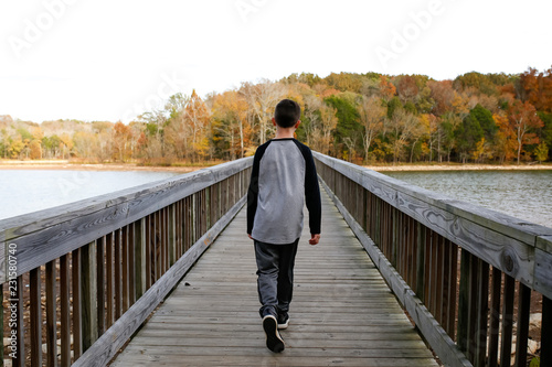 Fototapeta Naklejka Na Ścianę i Meble -  Boy Walking Across Bridge Over Lake In The Autumn