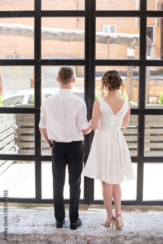 Charming bride and groom are holding hands on panoramic window background. Back view