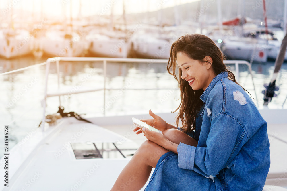 Fototapeta premium Young beautiful girl is sitting on the deck of a yacht with a smartphone