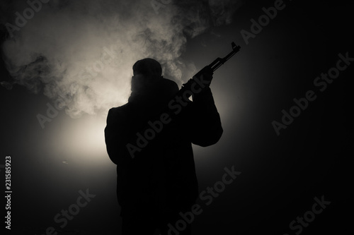 Silhouette of man with assault rifle ready to attack on dark toned foggy background or dangerous bandit in black wearing balaclava and holding gun in hand. Shooting terrorist with weapon theme decor photo
