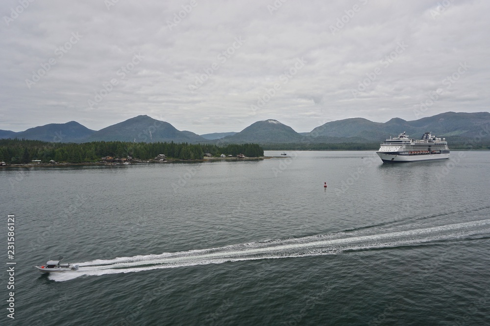 Under a cloudy sky, with mountains in the background, a speedboat races past a cruise ship in the Tongass Narrows, near Ketchikan, Alaska, USA.