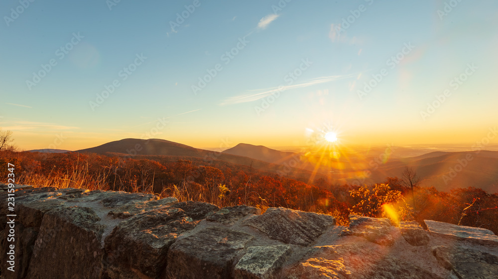 The sun appears on the horizon at daybreak highlighting the red, orange, and yellow autumnal colors of the trees in the forest at Shenandoah National Park during the fall