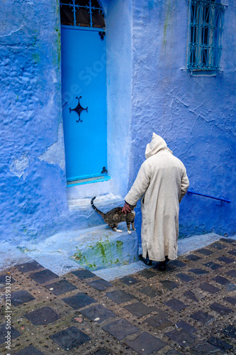 A Moroccan man wearing a traditional djellaba robe stops to pat a cat on a doorstep in the blue painted city of Chefchaouen in the Rif mountains of northern Morocco photo