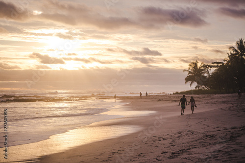 Couple walks holding hands along the long sandy paradise beach of Playa del Carmen, Santa Teresa Costa Rica during a colorful sunset